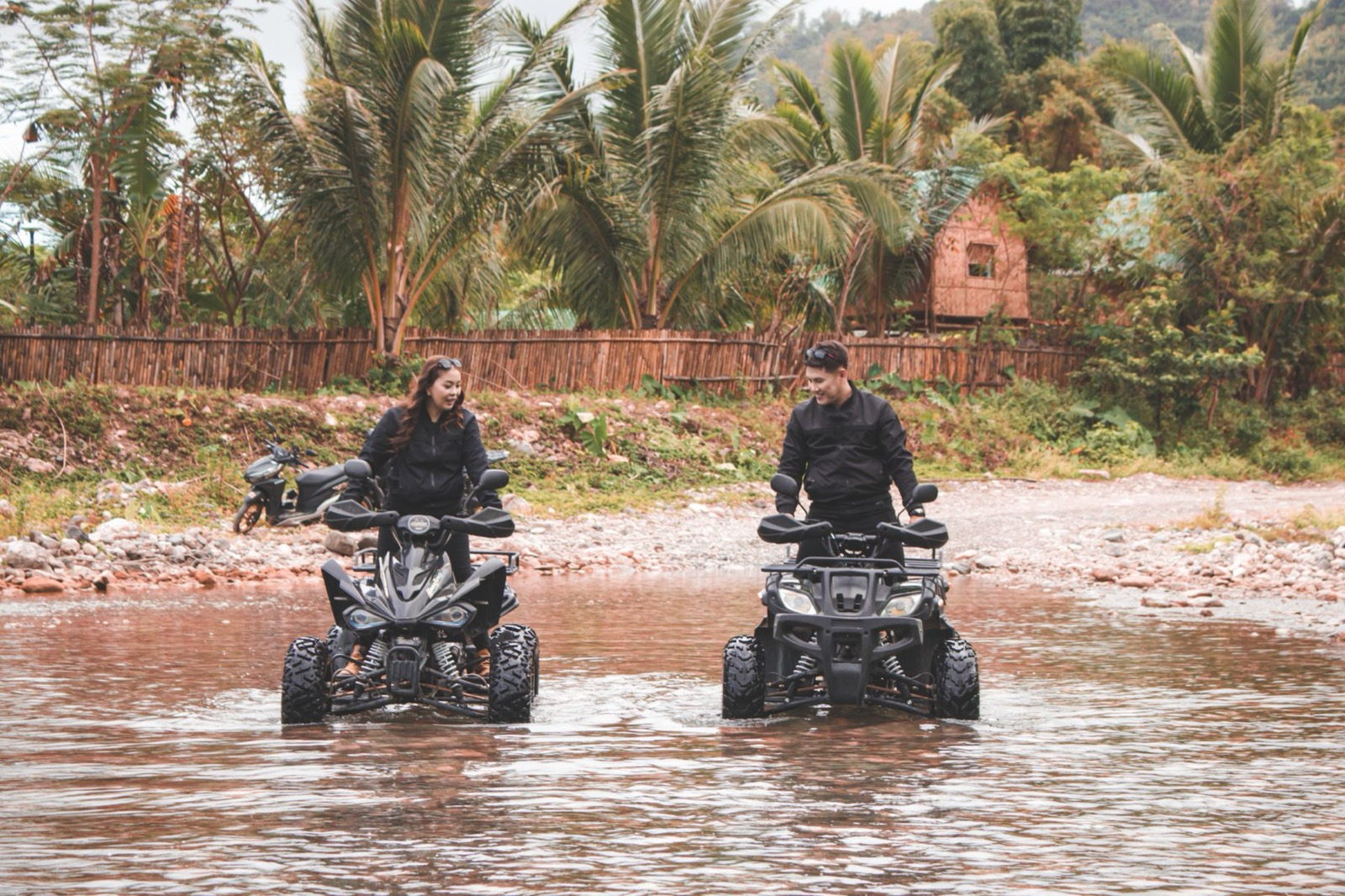 Two riders taking quad bikes through a shallow river between palms.
